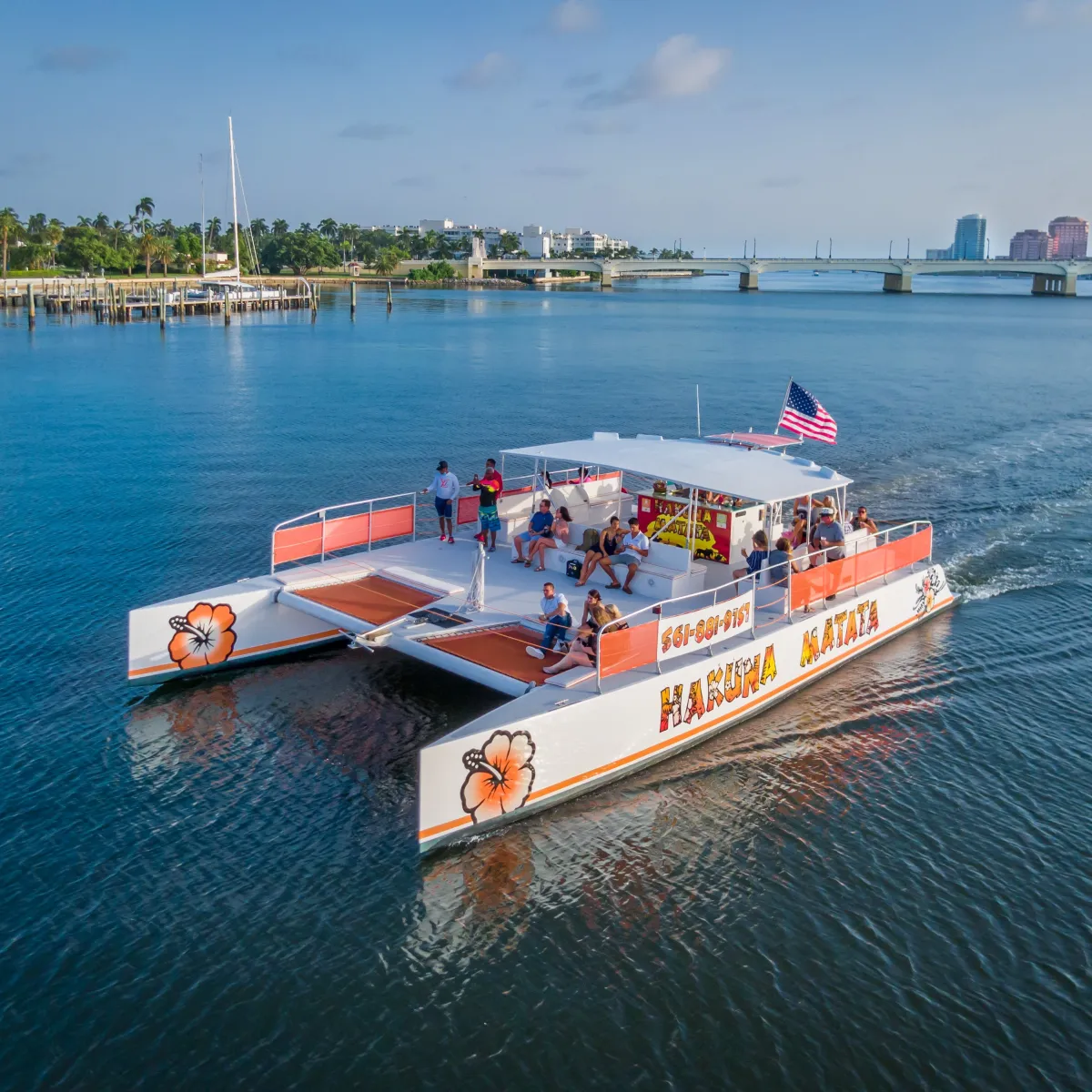 Tour catamaran with people, American flag, and hibiscus design cruising near a bridge.
