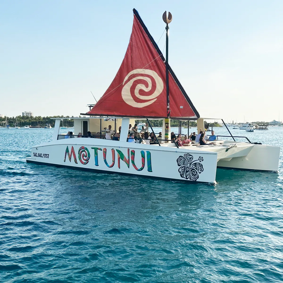 Catamaran with a red sail and the name 'Motunui' on water with people onboard.