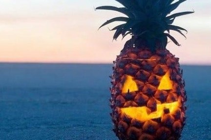 Pineapple carved like a jack-o'-lantern on a beach at sunset.