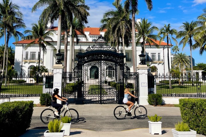 Two women biking past a large white mansion with a black wrought iron gate and palm trees.