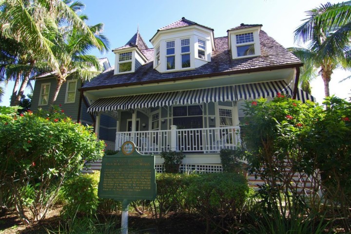 Historic house with striped awning, surrounded by palm trees and a garden.