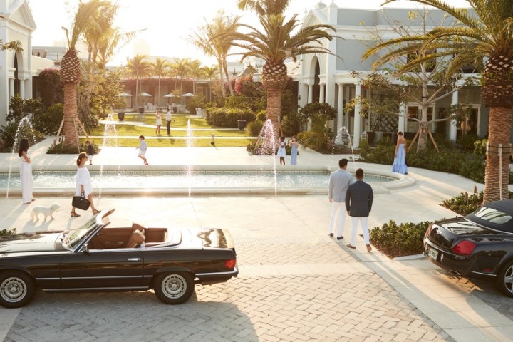 People in a luxurious courtyard with cars, palm trees, and fountains at sunset.