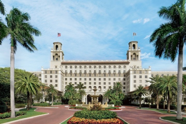 Large historic building with palm trees and a fountain in front, under a blue sky.