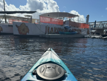 Kayak on water approaching docked boat with orange netting and sign.