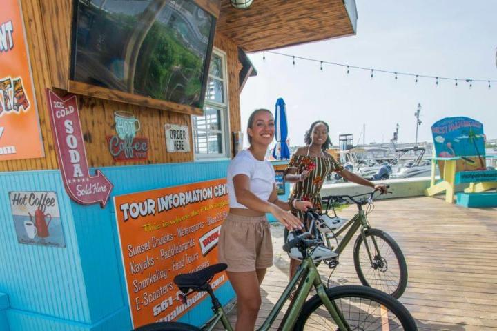 Two women smiling with bicycles near a tour information center by the waterfront.