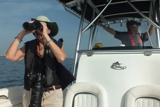 Two people on a boat; one uses binoculars, the other steers.