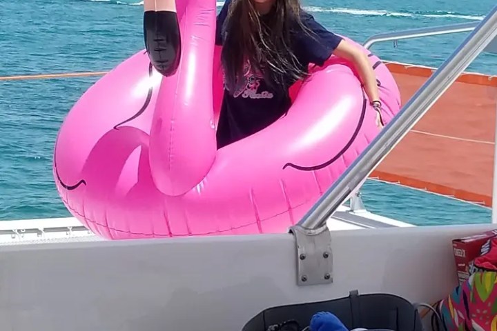 Person with long hair in a pink flamingo float on a boat with ocean in the background.