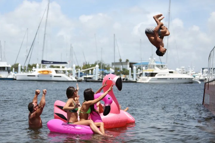 Man flips off pier into water, people on pink flamingo float watch, boats in background.