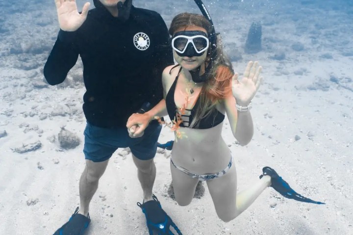 Two snorkelers underwater waving with clear masks and blue fins, sandy seabed visible.