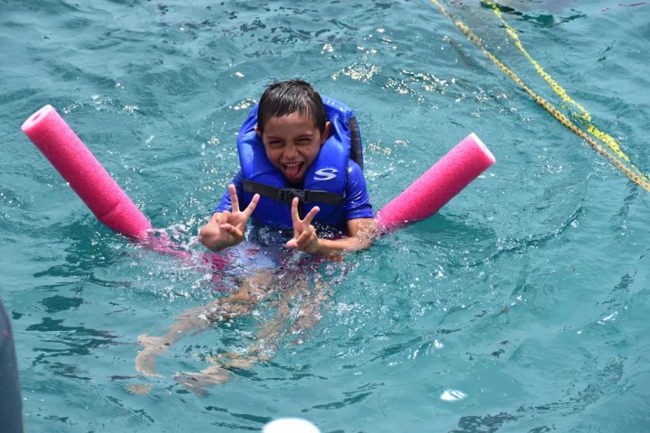 Child in blue life jacket swimming with pool noodles, making peace signs.
