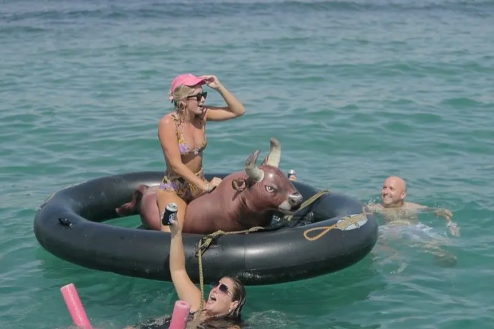 Woman on inflatable bull in ocean with friends swimming nearby, enjoying a sunny day.