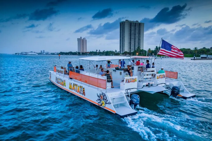 Catamaran 'Hakuna Matata' with passengers and American flag sailing near city skyline.