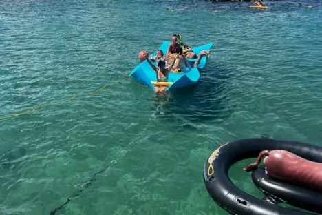 People in a pedal boat on clear blue water near a beach with palm trees and rocks.