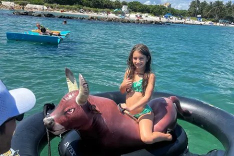 Girl on inflatable bull float in the ocean, sunny day with distant beach.
