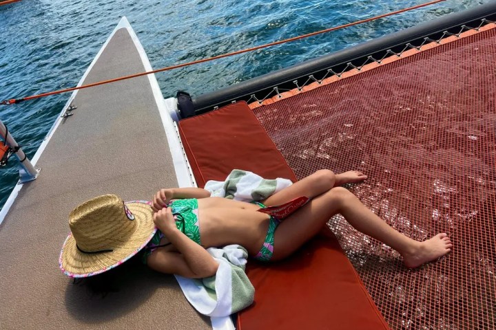 Child in swimsuit and hat relaxing on a boat's netting over water.