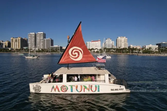 A sailboat with 'Motunui' painted on the side, sailing near a city skyline on a clear day.