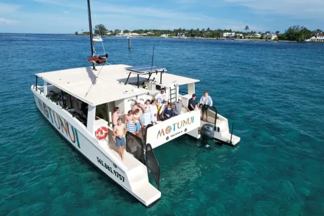 Boat named Motunui on clear blue water with eight people waving.