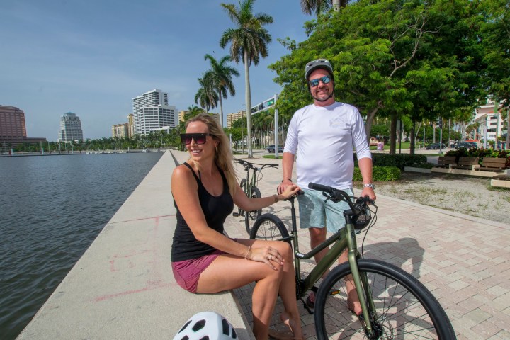 Two people with bikes by a waterfront path with palm trees and city buildings.