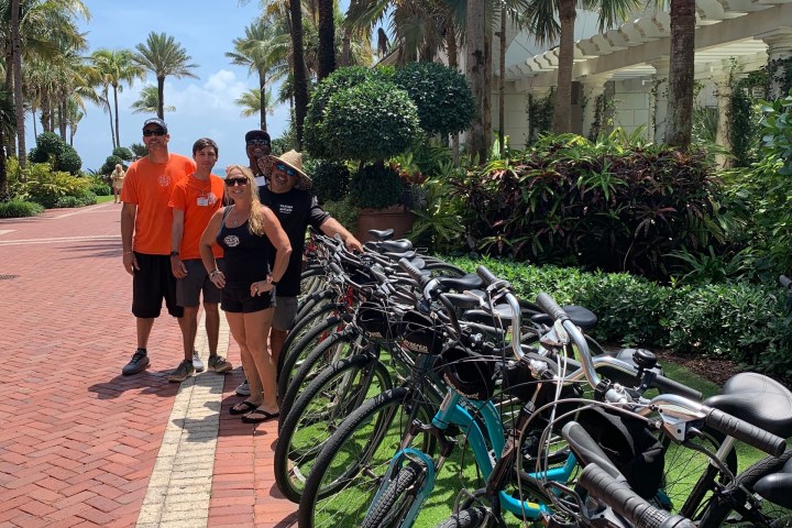 Group of people posing beside parked bicycles on a palm-lined path.