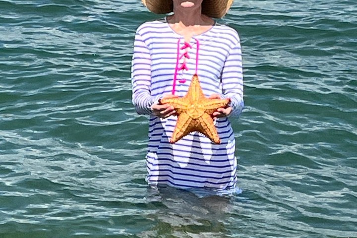 Person in striped dress holding a starfish in water, wearing a sun hat and sunglasses.