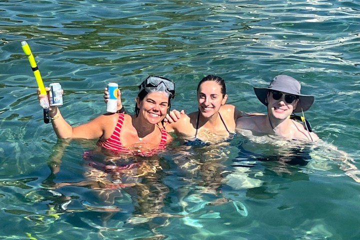 Three people smiling in water, one holding a drink can, wearing swim gear and hats.