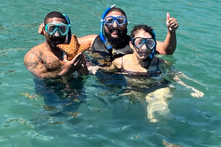 Three people snorkeling in clear water, one holding a starfish and giving a thumbs up.