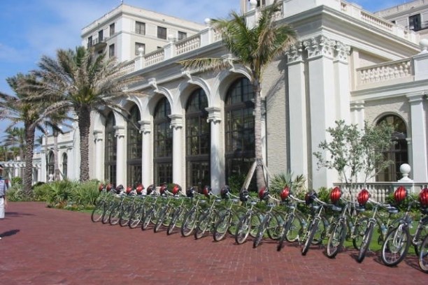 Row of bicycles with helmets in front of a large, white historic building with arches.